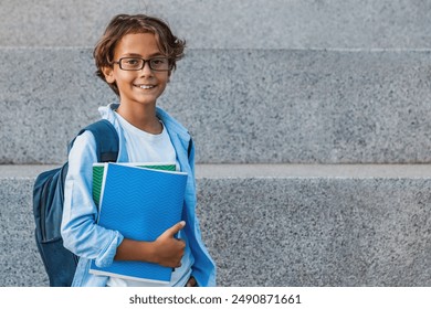 Portrait of happy caucasian young preteen boy elementary middle school pupil in eyeglasses with backpack holding notebook books outside the primary school. Education concept copy space - Powered by Shutterstock