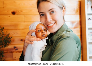 Portrait Of Happy Caucasian Young Woman Hugging Realistic Newborn African American Baby Doll And Looking At Camera, Indoors. Smiling Girl Holding Handmade Reborn Child Toy In Her Arms.