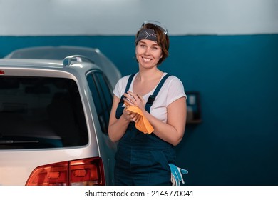 Portrait Of A Happy Caucasian Young Woman In A Blue Coveralls Waping Her Hands With A Rag. Car And Workshop In The Background. Work In An Auto Repair Shop.