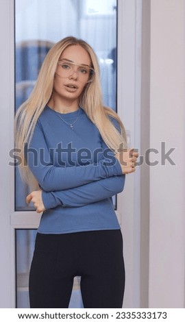 Portrait of happy charming cheerful blonde young girl in glasses, smiles, looking happy and glad, lively, with natural make-up, tanned, dressed in casual light blue long sleeve t-shirt