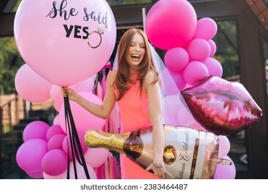 Portrait of happy charming redhead bride in summer wedding dress posing holding various colorful festive foil helium balloons standing by country house, closed eyes with happy expression.