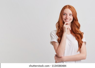 Portrait of happy charming redhead young woman with long wavy hair and freckles wears t shirt looks to the side, thinks and plans her birthday party isolated over white background
