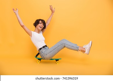 Portrait of a happy cheerful girl sitting on a skateboard with hands outstretched and looking at camera isolated over yellow background