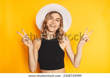 Portrait of a happy cheerful girl in white hat and black top showing peace gesture with two hands isolated over yellow background Victory peace gesture of happy curly blonde woman.
