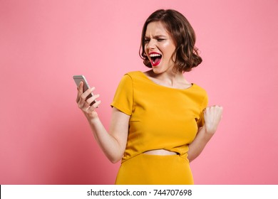 Portrait of a happy cheerful woman celebrating success while standing and looking at mobile phone isolated over pink background
