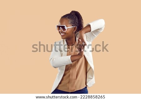 Portrait of a happy cheerful young black woman in a stylish jacket and party glasses smiling, dancing and having fun isolated on a solid beige colour studio background