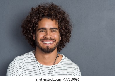 Portrait of a happy cheerful young man looking at camera. Handsome man with beard and curly hair standing against grey background. Close up face of multi ethnic young man isolated against grey wall.
