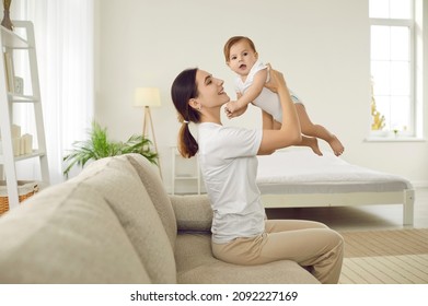 Portrait Of Happy Cheerful Young Woman Holding Her Baby And Playing With Her At Home. Side View Of Woman Sitting On Sofa In Bright Bedroom And Having Fun Lifting Asix Month Old Baby High In Air.