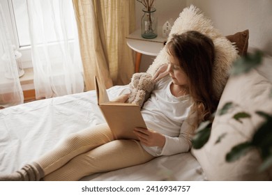 Portrait of happy child. Little beautiful curly girl hugs her plush rabbit and reads book in her bed. - Powered by Shutterstock