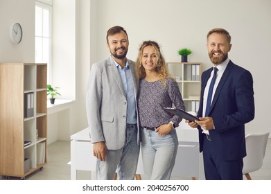 Portrait Of Happy Clients And Real Estate Agent. Young Married Couple Together With Realtor, Bank Specialist Or Mortgage Broker Standing In Office, Smiling And Looking At Camera