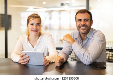 Portrait of happy colleagues sitting at cafe during break. Young Caucasian businesswoman with pc tablet and mid adult businessman sitting at table, looking at camera and smiling. Colleagues concept