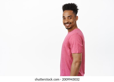 Portrait of happy and confident african american guy, standing in profile, turn head at camera with big cheerful smile onhis face, standing in pink t-shirt against white background