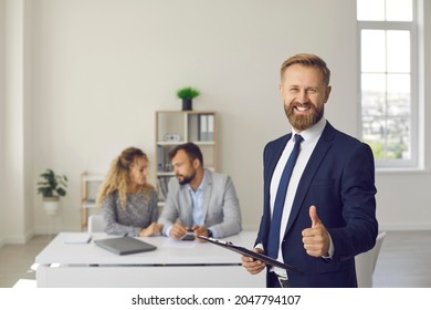 Portrait Of Happy Confident Male Real Estate Agent Or Mortgage Broker In Suit Looking At Camera, Smiling, Offering Best Deal And Giving Thumbs Up Standing In Office With His Clients In Background