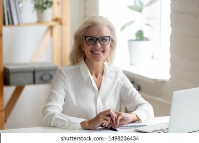 Portrait of happy confident senior grey-haired businesswoman in spectacles sit at office desk look at camera smiling, overjoyed glad aged woman worker wear glasses posing making picture at workplace