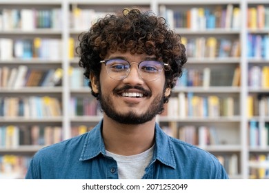 Portrait Of Happy Confident Smiling Young Indian Male Student Or Librarian Looking At Camera Standing Over Library Book Rack On Blurred Background. Education And Knowledge Information Concept