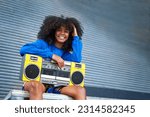 Portrait happy, cool young woman with curly hair holding yellow ghetto blaster at corrugated metal wall in alley