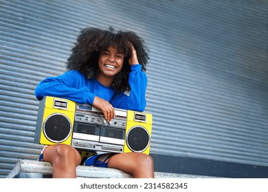 Portrait happy, cool young woman with curly hair holding yellow ghetto blaster at corrugated metal wall in alley