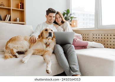 Portrait of happy couple hugging dog, golden retriever, selective focus using laptop online shopping, sitting on sofa at home. Smiling girlfriend and boyfriend watching video. Technology concept - Powered by Shutterstock