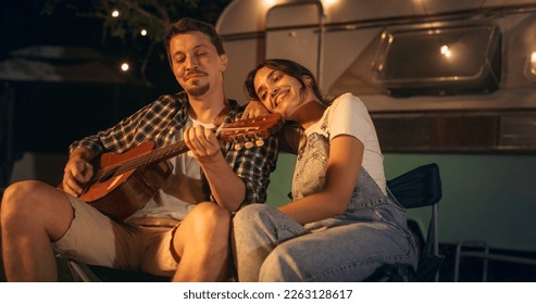 Retrato de una pareja feliz pasando tiempo en una fogata, tocando música de guitarra y cantando canciones divertidas en un área de Motorhome Camping Rest. Los socios encantadores disfrutan del verano y viajan juntos