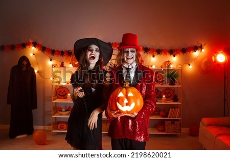 Portrait of happy couple in spooky costumes with Jack-o-lantern. Man in red suit and top hat and young lady dressed as witch standing together and laughing in living room decorated for Halloween party