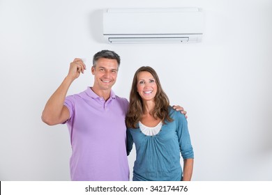 Portrait Of Happy Couple Standing In Front Of Air Conditioner With Remote In Man's Hand
