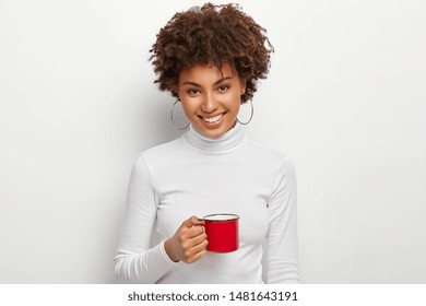 Portrait of happy curly haired woman with toothy smile, holds red mug of hot drink, looks straightly at camera, being coffee lover, dressed in white turtleneck jumper, has spare time, break after work - Powered by Shutterstock