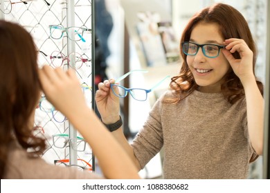 Portrait Of A Happy Cute Girl Trying On New Glasses Looking In The Mirror At The Optometrists Store. Adorable Child Wearing Glasses At The Opticians Shop Copy Space