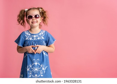 portrait of a happy cute little girl making a heart gesture, isolated on a pink studio background. Smiling baby a child with wavy hair 5 years old a charming contented cutie shows a sign of love.