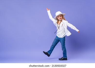 Portrait of happy cute little girl in a hat and unisex clothes on a purple background in full growth. stylish children's model is having fun and feels stunned, amazed. Advertising of children's goods
