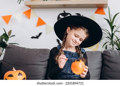 Portrait happy cute little kid girl in black hat  painting small orange pumpkin sit cozy couch in living room at home  Child celebrating traditional festival halloween thanksgiving concept