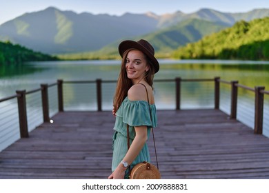 Portrait of happy cute smiling attractive traveler girl wearing hat and green dress standing alone on pier with lake and mountains view. Enjoying serene quiet peaceful atmosphere in nature