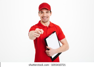 Portrait of happy delivery man in red t-shirt and cap smiling and pointing finger on camera while holding clipboard isolated over white background