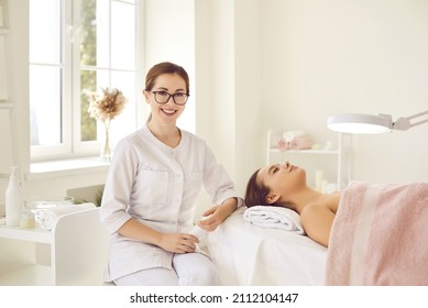 Portrait Of Happy Dermatologist, Skin Therapist, Beautician And Skincare Professional In Workplace. Smiling Beautiful Young Woman In White Uniform And Glasses Sitting Next To Client On Procedure Table