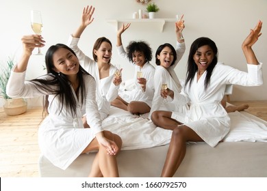 Portrait Happy Diverse Girls Wearing White Bathrobes Holding Champagne Glasses, Sitting On Bed, Looking At Camera, Beautiful Young Women Celebrating Birthday Or Wedding In Spa Or Hotel, Hen Party