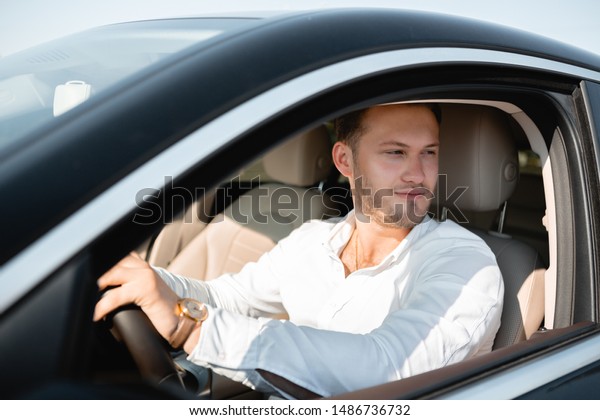 Portrait of a happy driver in a white shirt\
driving his luxary car. Concept of\
success.