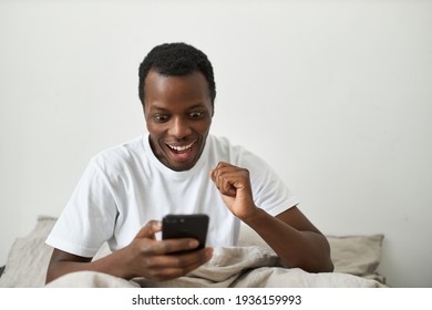 Portrait Of Happy Ecstatic Young Afro American Male Investor Making Money In Stocks Using Online Banking Application On Mobile Phone, Having Excited Facial Expression, Cheering, Opening Mouth Widely