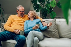 Portrait Of A Happy Elderly Couple Hugging And Relaxing Together On The Sofa At Home And Drinking Coffee. Senior Couple Having A Conversation Together While Relaxing On The Couch. Copy Space.