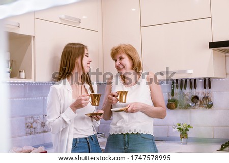 Portrait of a happy elderly mother and daughter in modern light kitchen interior.Women hold golden cups with coffee,tea in hands laughing and looking on each other.Tender relationship in family.Text