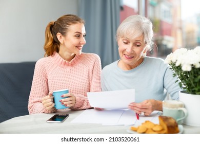 Portrait Of Happy Elderly Woman Sitting With Adult Daughter At Home Table With Cup Of Tea, Reading Letter Together .