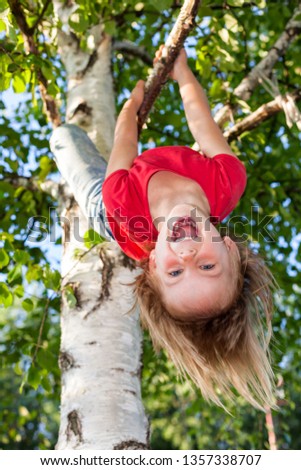 Portrait of happy elementary age girl wearing red tee shirt hanging upside down from a tree branch - summer fun concept