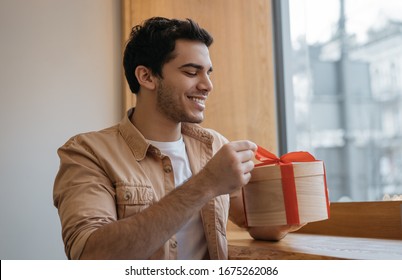 Portrait Of Happy Emotional Man Opening Gift Box. Young Attractive Indian Guy Holding Birthday Present And Smiling, Sitting At Home	