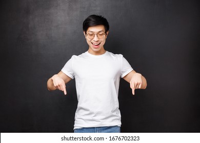Portrait of happy enthusiastic asian man in sunglasses and t-shirt, looking and pointing down as watching something interesting happening at bottom promo banner, black background