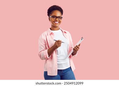 Portrait of happy entrepreneur or business assistant. Attractive African American woman in shirt, tee and glasses standing on pink background, holding pen and clipboard, looking at camera and smiling - Powered by Shutterstock