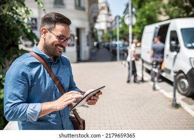 Portrait Of Happy Entrepreneur Holding Digital Tablet And Looking At The Screen. Young Handsome Businessman Carrying Laptop Bag. He Is Standing On Sidewalk In The City.