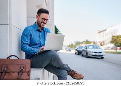 Portrait Of Happy Entrepreneur Holding And Working On Laptop While Sitting At The Urban Street. Young Handsome Businessman Carrying Laptop Bag And Doing Business Outside In The City Street.