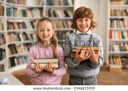 Portrait of happy european boy and girl pupils holding a stack of books, standing in library and smiling at camera. Children holding heap of books