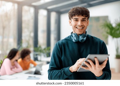Retrato de un estudiante europeo feliz que sostiene una tableta digital para una lección de video o una aplicación educativa, que estudia con compañeros de clase en el salón de clases, mujeres sentadas en la mesa sobre el fondo