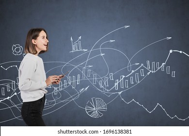 Portrait Of Happy European Teen Girl With Smartphone Standing Near Blackboard With Blurry Financial Graphs Drawn On It. Concept Of Business Education And Stock Market.