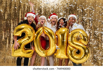 Portrait of a happy excited multhiethnic group of friends in red hats celebrating New Year while standing and holding air balloon golden 2018 numbers isolated over golden shiny background - Shutterstock ID 744940000