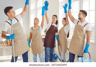 Portrait of happy excited smiling young team of janitors in uniform from professional cleaning service standing in a circle with hands up finishing work in clients apartment. Housework concept. - Powered by Shutterstock